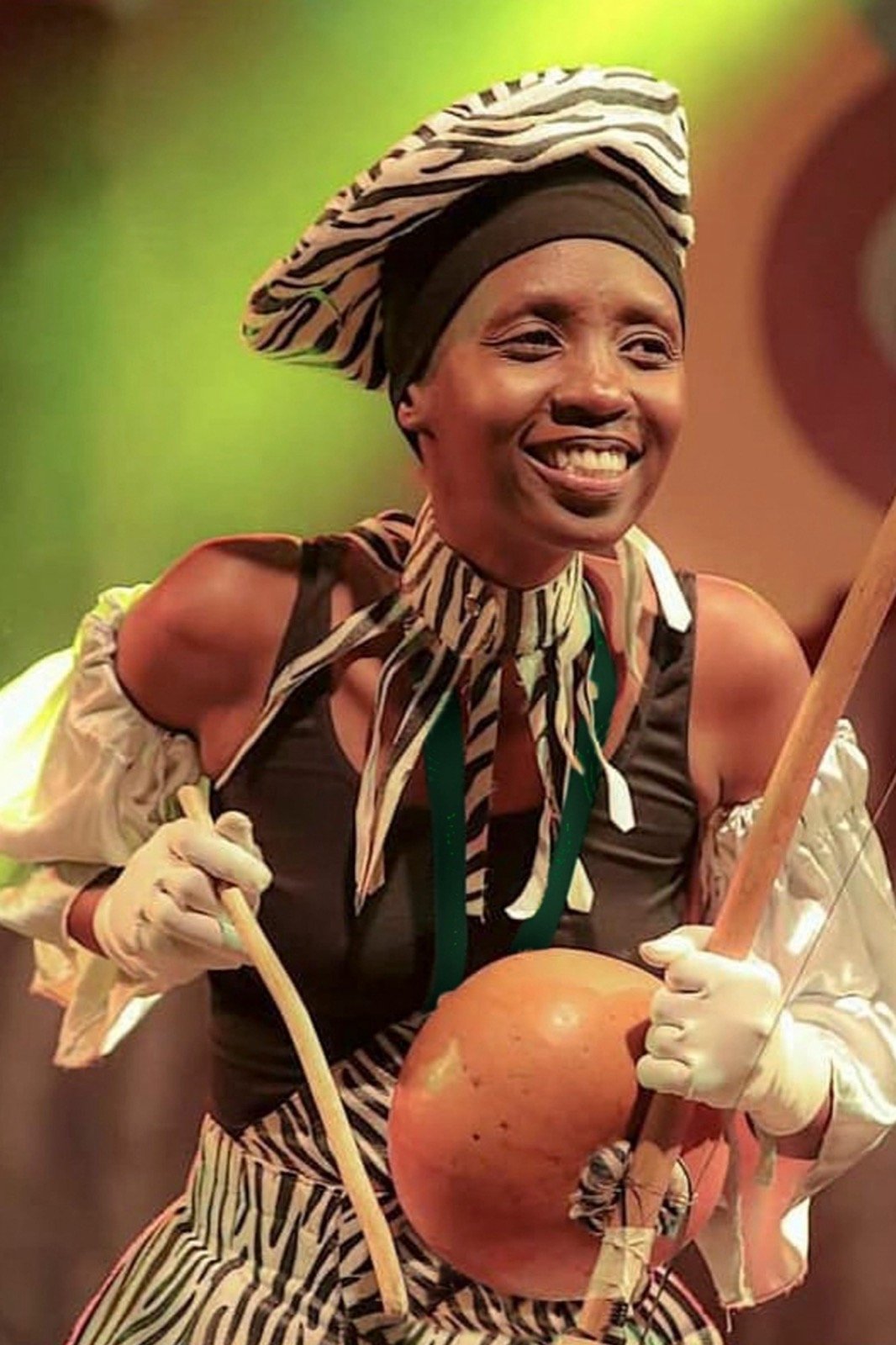 Bernice the Bell sur scène, souriante, coiffe zébrée, jouant d'un arc musical à calebasse